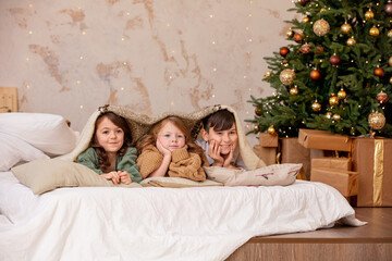 Three children, two girls and a boy, happy together at home are lying on the bed under the blanket by the decorated Christmas tree