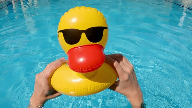 Point of view of person two hands holding a yellow inflatable duck on surface of water outdoors in summer. Children hands gaming with cute toy in blue swimming pool transparent water.