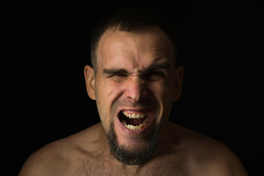 Bearded man screaming with his eyes squinted. Portrait of a screaming Caucasian man without a shirt on a black background. Life problems.