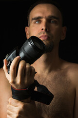 Breast portrait of photographer on black background. Shirtless man holding camera with one hand near his face. Low key.