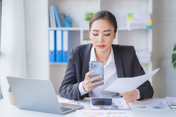 Young beautiful asian woman using smartphone and working with laptop while sitting at office desk, working from home concept.
