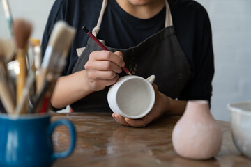 Craftswoman creates new masterpiece in work studio making sketch of future pattern on white mug. Lady master wants to decorate kitchen with painted clay dish using pencil to draw interesting pattern