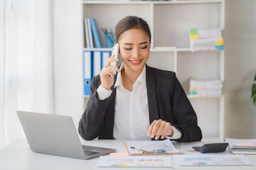 Young beautiful asian woman using smartphone and working with laptop while sitting at office desk, working from home concept.