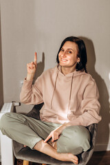 Obraz premium A girl in a pink hoodie sits on a gray chair near the window, radiator and shows with a finger that an idea has come. Front view