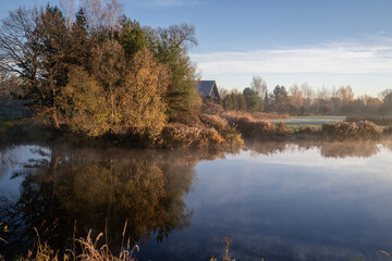 beautiful misty morning over river Vircava near Jelgava town in Latvia. Smooth calm water surface. Golden autumn morning sunrise light. Trees yellow leaves, reflection of river bank flora  blue sky 