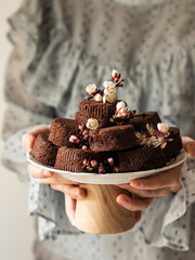 A young Caucasian woman holding in her hand's cake stand with cocoa round brownies and some flower blossoms