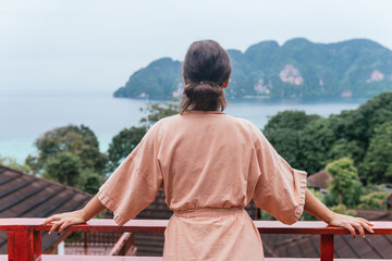 Fototapeta premium Unrecognizable woman in a bathrobe leaning on the railing of her terrace while looking at the views of the sea and the mountains in front of her.