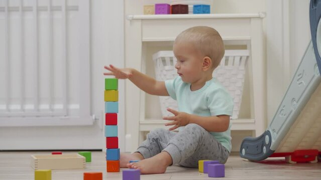 Cute little toddler baby child playing with wooden building blocks on the floor at home. Sweet happy boy builds a tower of colored wood blocks for toddlers.
