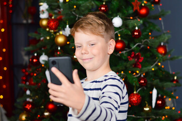 Christmas eve - smiling boy talking photo of themselves on mobile phone. Christmas tree with lights on dark red as background.