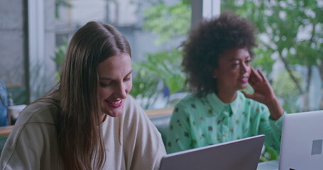 Two diverse young women at coffee shop in front of laptop computers. Female work colleagues working remotely