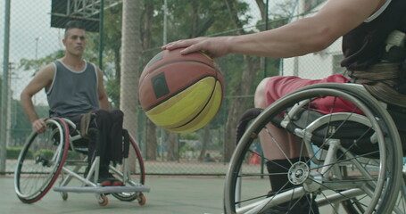 Obraz premium A paraplegic male athlete receiving ball from colleague outdoors. Two disabled athletes playing basketball in sport court outside. Sport and disability concept