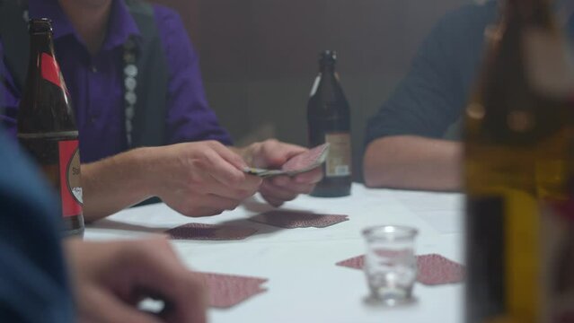 Close up of a group of men playing a tyrolean card game with hands distributing cards to other players with a bottle of beer in the foreground in a hazy room. Shot in 4K