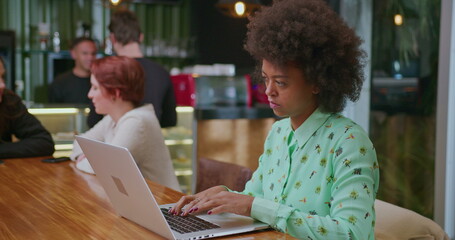 One focused African American young woman in front of laptop seated at coffee shop. A concentrated black female person working remotely at cafe place interior