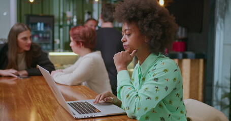 Fototapeta premium One focused African American young woman in front of laptop seated at coffee shop. A concentrated black female person working remotely at cafe place interior