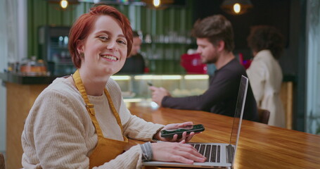 One happy female barista seated at coffee shop table holding phone in front of laptop computer turns head to camera smiling