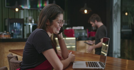 Female hispanic barista employee working in front of laptop computer taking online delivery. Young woman using technology speaking with client and multi tasking