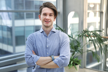 Portrait of handsome happy positive European young student or office worker employee in shirt standing indoors with his hands crossed looking at camera, smile. University or college.