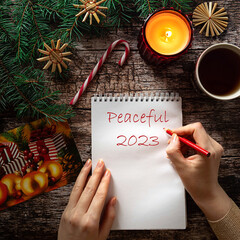 Woman's hand writing Peaceful 2023 on a notepad sheet in winter holiday setting