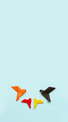 four paper origami pigeons black, orange, red and yellow on light blue background, vertical, 16:9