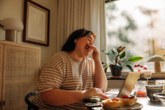 Cheerful young woman covering mouth with hands while sitting at table by window