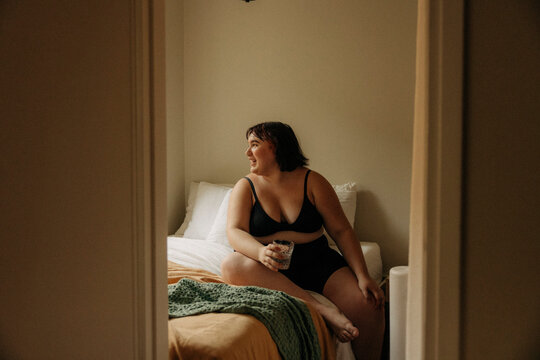 Full length of happy young woman wearing lingerie sitting with drinking glass in bedroom