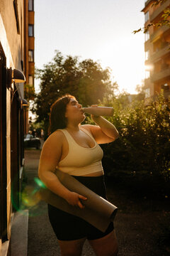 Young woman with exercise mat drinking water from bottle at back yard