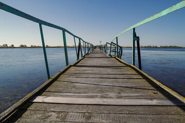 Obraz premium Wooden bridge over river landscape photo. Beautiful nature scenery photography with wide watercourse on background. Idyllic scene. High quality picture for wallpaper, travel blog, magazine, article