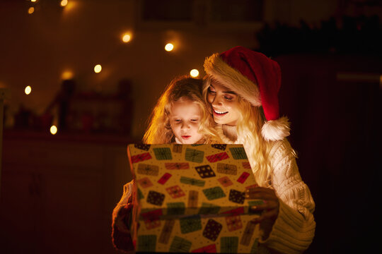 Front view of blonde mother wearing christmas hat opening present, gift box with surprised daughter. Happy family celebrating holiday, smiling. Concept of new year and christmas.