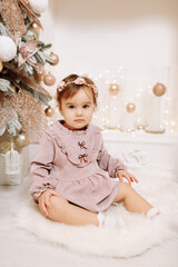 Fototapeta premium Adorable baby girl sitting by the festive decoration fireplace under fir tree. Child in soft pink dress with many gift boxes presents. Happy Holidays, New year. Cozy warm winter evening at home.