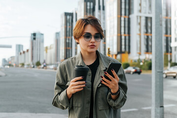 Portrait of beautiful stylish young asian woman with short haircut and glasses holding coffee and using smartphone, looking at mobile phone screen while standing on city street