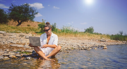 Young woman freelancer traveler wearing white shirt anywhere working online outdoors using laptop enjoying lake view. Happy female downshifter in sunglasses holding computer at sea coastline 