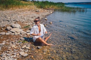 Young woman freelancer traveler wearing white shirt anywhere working online outdoors using laptop enjoying lake view. Happy female downshifter in sunglasses holding computer at sea coastline 