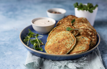 Fototapeta na wymiar Vegetable fritters or pancakes made of zucchini, broccoli or spinach on plate with microgreen and dip, blue background. Healthy vegetarian food.