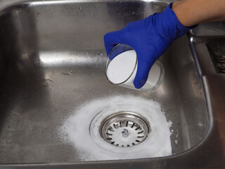 Gloved hand putting baking soda on drain In sink from glass jar - obrazy, fototapety, plakaty