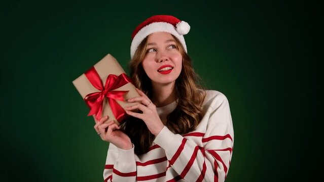 Woman in santa hat shaking new year gift trying to guess what's inside isolated on green background. Smiling girl in sweater and christmas box