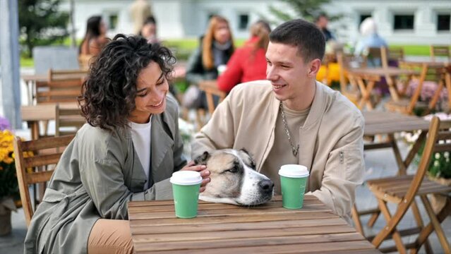 A happy couple outdoors near a cafe. Petting their dog, coffee on the table. Autumn atmosphere. Slow motion