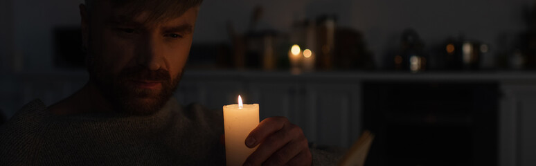 man holding burning candle in dark kitchen during electricity outage, banner.