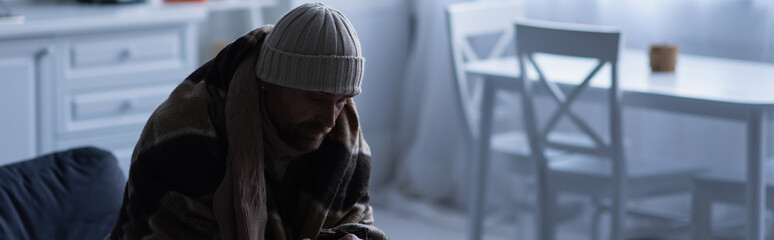 frozen man sitting in warm blanket and knitted hat at home in twilight, banner.