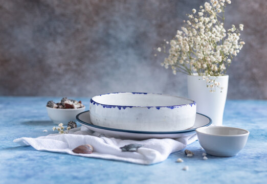 Handmade ceramic white plates with blue stripe on the edge, dried flowers and empty seashells on blue concrete background.