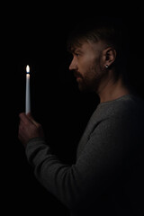 side view of adult man holding burning candle in darkness isolated on black.