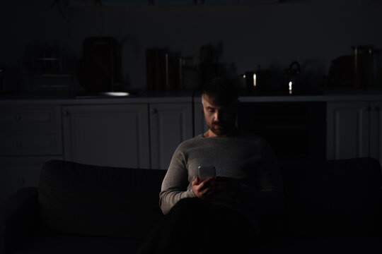man sitting with mobile phone in dark kitchen during energy blackout.