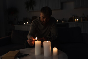 man lighting candles near book and mobile phone while sitting in kitchen during energy outage.