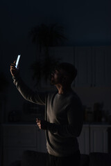 man standing in dark kitchen with smartphone in raised hand and searching for connection during...