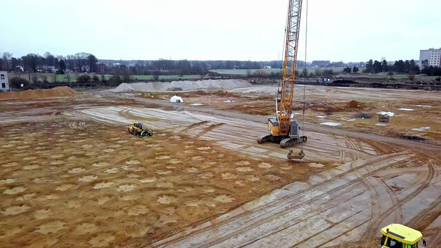 Dynamic soil compaction - a tall crane drops a heavy weight on the foundation of a huge construction site - aerial pull back view 