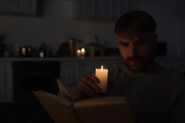 man holding lit candle while reading book in dark kitchen during electricity shutdown.