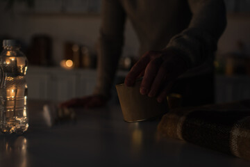 cropped view of man holding canned food near warm blanket and bottled water in dark kitchen.