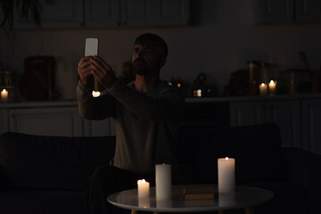 man sitting near burning candles in dark kitchen and catching mobile connection on smartphone.