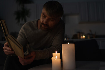 man reading book while sitting in dark kitchen near burning candles.
