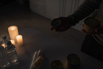 partial view of man holding cans near table with bottled water and candles in dark kitchen.