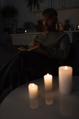 man reading book during electricity shutdown near lit candles in dark kitchen.